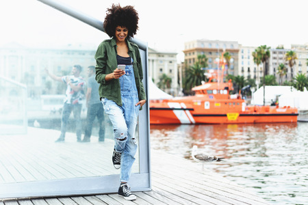 a beautiful African-American girl dressed in casual clothes reads a test message on smartphone while relaxing on the waterfront.の写真素材