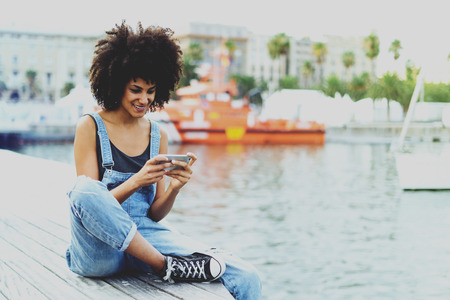 beautiful black girl with amazing afro hair checks mail through cellphone in rest time on embankment warm summer evening.young casual clothes,mixed race girl watching video on mobile phone and smilingの写真素材