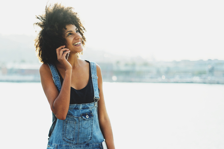 Happy female with afro hair and casual outfit talking on phone walking along the sea.の写真素材