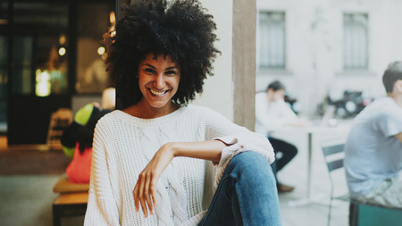 Half length portrait of a cheerful mulatto female with afro hair style wearing stylish clothes smiling at the camera while sitting in a modern city coffee shop on a weekend day on a blurred backgrond.の写真素材