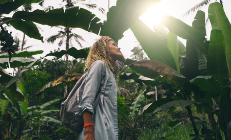 View from the bottom photo of a beautiful caucasian female with curly hair standing amid the huge green leaves and impressed with wild tropical nature during journey through the jungles.の写真素材
