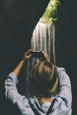 View from the back photo of blonde girl taking photo of an outstanding beautiful waterfall by the camera of her cellular phone in the depth of tropical jungles during summer vacation. の写真素材