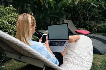 A hipster girl is relaxing at pool during vacation and chatting with friends by mobile phone and laptop. Young designer is sitting with a smartphone and a portable computer with blank displays.の写真素材