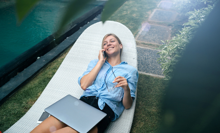 Young blonde female is working on laptop and smartphone beside the pool on luxury tropical villaの写真素材