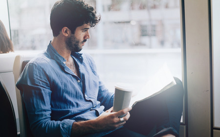 Young bearded office employee male reading newspaper and drinking takeaway coffee while going to the work by public transport to avoid morning traffic jam for cars.の写真素材
