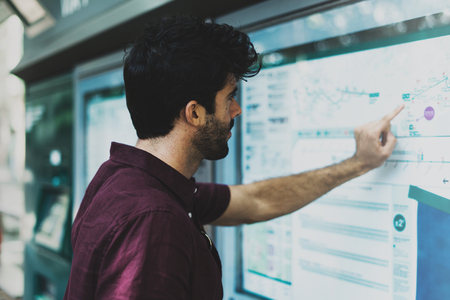 Young bearded tourist man searching the route on a public transportation system map in unknown city.