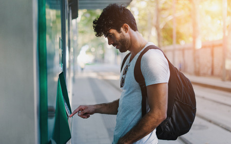Bearded handsome hipster guy wearing casual clothes with backpack is buying bus tickets at self...