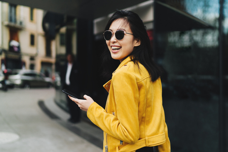 Photo in motion of a smiling asian woman with long dark hair in yellow leather jacket holding a mobile phone while walking the street. Cheerful student girl chatting online by a smartphoneの写真素材