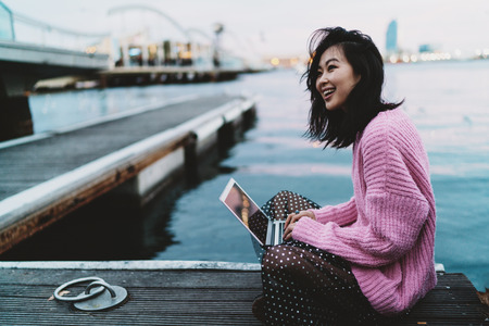 Charming asian hipster girl surfing the web on a portable computer while sitting at the city berth. Successful fashion blogger writing emails to the clients by a laptop connected to wi-fi.の写真素材