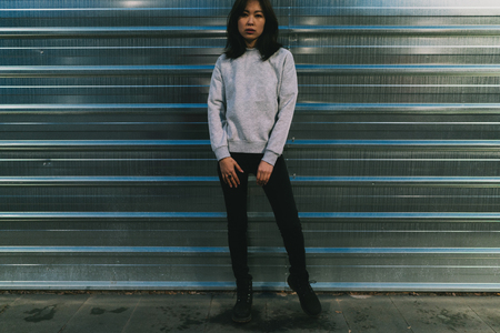 Stylish asian hipster girl wearing blank light gray sweatshirt and black jeans standing against urban metallic background with copy space area for your text message or advertising content.の写真素材