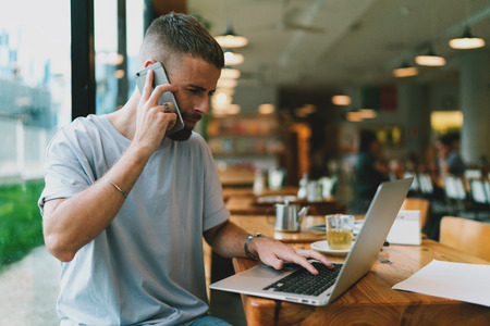 Serious male architect is talking by mobile phone and looking at laptop display while sitting at modern coffee shop. Bearded man spending free time while having conversation on cell phone.の写真素材