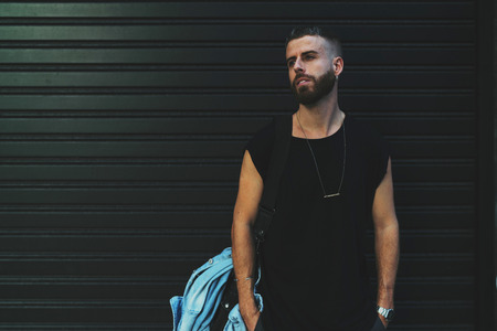 Portrait of a serious young bearded male hipster standing on a dark wall background. Handsome male wearing black blank t-shirt looking aside. Empty space on a t-shirt for your logo or design.の写真素材