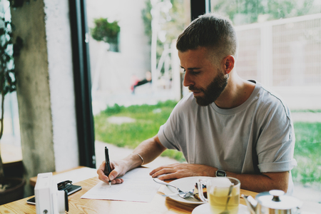Creative web illustrator is making sketches for a new project while enjoying relaxing herbal tea in a cafe. Skilled casually dressed student making notes in notepad for the diploma project.の写真素材