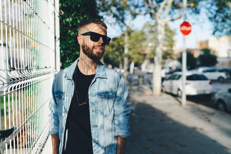 Half-length portrait of young stylish bearded hipster guy in sunglasses wearing black t-shirt and a light blue jeans enjoying the summer day while walking the city on a weekend. の写真素材