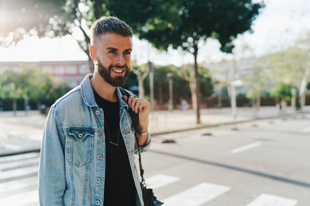Smiling handsome hipster guy wearing light blue jeans jacket crossing a street in good mood....