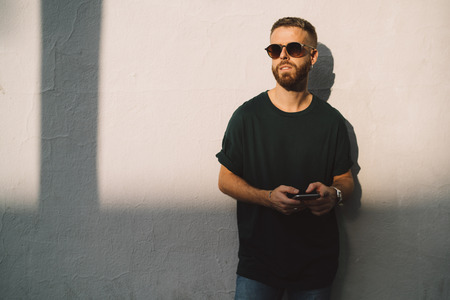 Half-length portrait of bearded make guy texting to the friend by a mobile phone while standing outdoors. Stylish student guy surfing the web on a smarthone while standing against white concrete wall.の写真素材