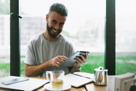 International student smiling at the camera while doing homework by using a tablet computer and other devices. Successful freelancer reading emails online while having tea break in a cafe.の写真素材