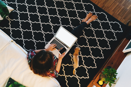 View from the top photo of young female sitting on a carpet with a laptop on her knees with blank white display for your content. Student girl studying online from home with a dog beside her.の写真素材