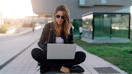 Beautiful caucasian female is sitting at city area with a laptop connected to wi-fi. Young entrepreneur is working on a portable computer while sitting outdoors on a blurred urban background.の写真素材