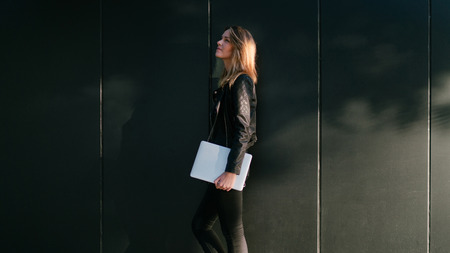 Stylish young woman wearing black jeans and leather jacket is looking aside while standing with a laptop in her hand beside the black wall.Caucasian female with portable computer on a wall background.の写真素材