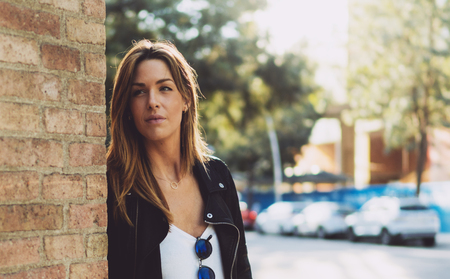 Half-length portrait of young stylish caucasian woman with long hair wearing leather jacket looking aside while standing on a street leaning on a brick wall. Pensive attractive female looking aside.の写真素材