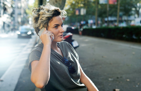 Hipster girl is talking to the friends by a mobile phone while sitting on the bench on empty evening...