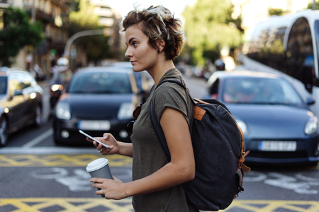 Happy young woman with blonde short hair is messaging to the friend by a smartphone while crossing the road with a blank paper cup in her hand. Attractive tomboy girl is reading sms on mobile phone.の写真素材