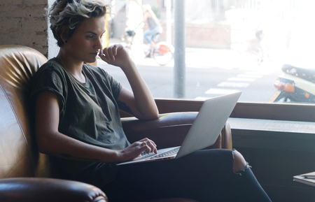 Blonde hipster girl is reading emails on a screen of a modern portable computer while sitting in a cafe.の写真素材