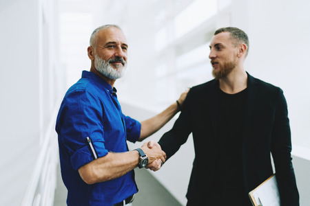 Portrait of a smiling manager shaking hands with his new subordinate employee while standing in office space interior. Caucasian male entrepreneur congratulates his partner with new project launch.の写真素材