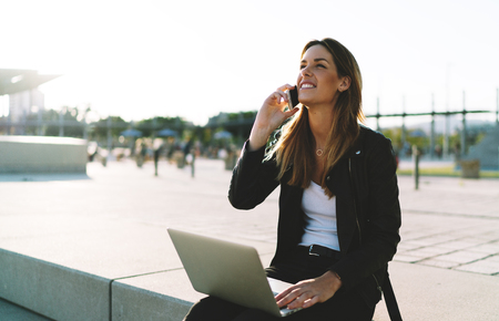 Young entrepreneur female happy to receive good news by a mobile phone from her partners. Stylish hipster girl cofirming by a smartphone call a flight booking she made online on a laptop.の写真素材