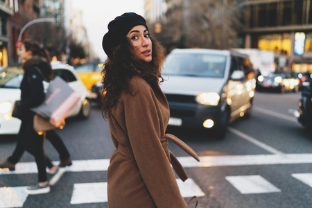 Portrait of a charming caucasian woman with beautiful long dark curly hair wearing coat and a beret looking back at the camera while crossing the street on a blurred urban background.の写真素材