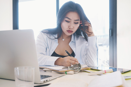 Young business woman is making business calls by smartphone from the home office. Professional designer is sitting at the table with a laptop on it. Young asian lady is having a talk by a mobile phoneの写真素材