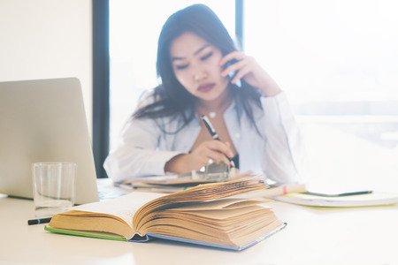 Young entrepreneur is making business calls by smartphone from the home office. Freelancer specialist is sitting at the table with a laptop on it. Young asian woman is having a talk by a mobile phoneの写真素材