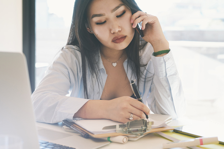 SMM specialist is making business calls by smartphone from the home office. Beautiful model look asian woman is sitting at the table with a laptop on it while talking by a cellphone.の写真素材