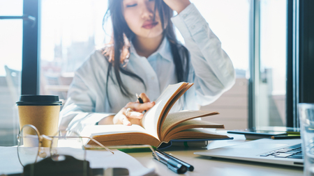 Young entrepreneur female is working at home on a sunny day. Beauty blogger is preparing new article on a rerto style clothes. Asian woman is sitting at the table with a book and laptop on it.の写真素材