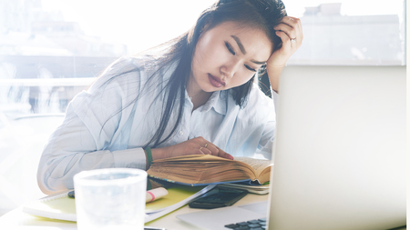 High school student girl is reading textbook while sitting beside the big window on a sunny day. Young attractive asian girl is searching information while preparing to the new presentation.の写真素材