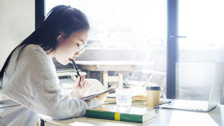 Young freelancer female is reading book while sitting beside the big window on a sunny day. Young attractive asian female is searching information while preparing to the new presentation.の写真素材