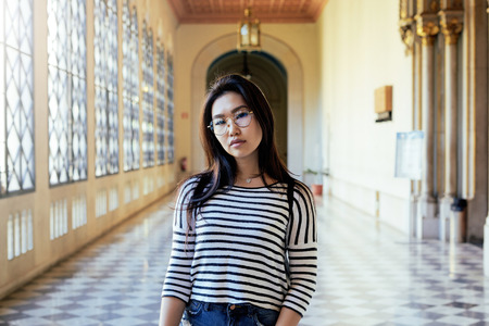 Asian student girl wearing glasses is looking at the camera while standing on the vintage hall background. A hipster female wearing casual clothes is walking the old city historic building.の写真素材