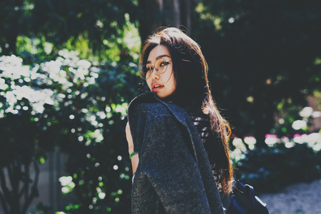 Portrait in motion of a beautiful asian woman with long dark hair wearing glasses with a jacket on her shoulder turning back and looking at the camera while walking the national park on a sunny day.の写真素材