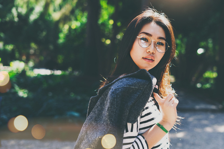 Charming brunette female in glasses looking at the camera while standing outdoors on the natural background at the city park. Model look asian woman relaxing at the city garden on a sunny day.の写真素材