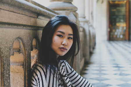 Beautiful asian female with long hair wearing casual outfit looking at the camera while sitting on a floor at the college area. Chinese tourist woman relaxing at the city museum during weekend journeyの写真素材