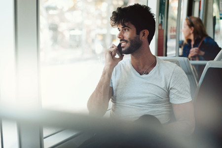 Young freelancer guy is having business call on a smartphone connected to public wi-fi while sitting in a city tramway. Attractive model look bearded man is chatting with friends by a mobile phone.の写真素材