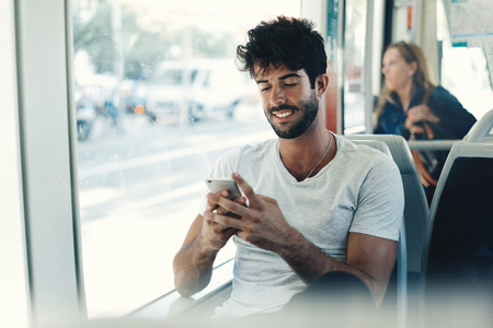 Bearded hipster guy is reading emails on a display of a smartphone connected to public wi-fi while sitting in a city bus. Handsome male is looking at the screen of a mobile phone while texting.の写真素材
