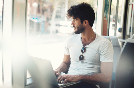 Bearded hipster guy is working on a portable computer connected to public wi-fi while sitting in a city bus. Young attractive male is looking at the window while typing messages by a laptop.の写真素材