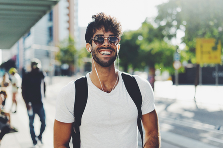 Young travel blogger is ready for a new weekend trip. Bearded backpacker is standing on a blurred station background. Student is relaxing on a weekend. Hipster guy wearing sunglasses smiling at cameraの写真素材