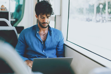 Young man is reading emails on portable computer while sitting in a city bus beside a window. Hipster guy with dark curly hair wearing light blue shirt is going to the office by a public transport.の写真素材