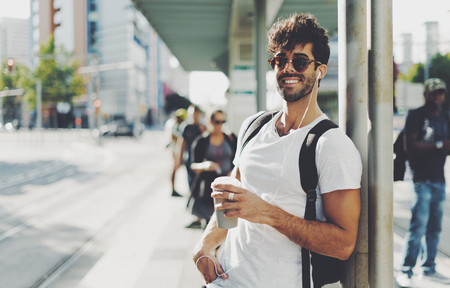 Handsome guy with dark curly hair wearing sunglasses is smiling at the camera while standing on the street with a cup of coffee on a sunny day. Bearded male is waiting for a train on a station.の写真素材