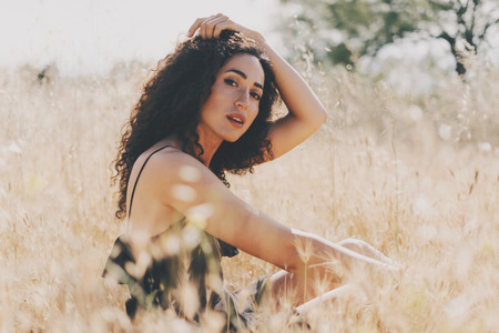 Cute caucasian female with long dark curly hair is looking at the camera while sitting amidst the field. A model look woman in a black summer dress is relaxing outdoors  on a sunny day.の写真素材