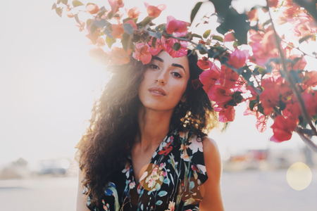 Young model look female wearing colorful summer dress is looking at the camera while standing beside the flowers on a summer day. A brunette hipster girl with long curly hair is having rest outdoors.の写真素材