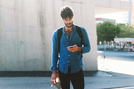 a young attractive usually dressed hipster guy with a backpack uses a e-map on modern mobile phone and holds coffee cup. a handsome guy is using a smartphone to connect to a public network wifiの写真素材